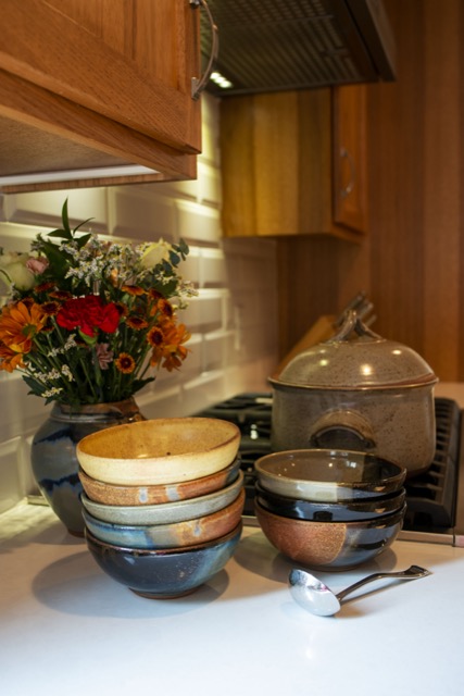 a vertical framed picture. two stacks of clay coyote chili bowls, a soup ladle and a clay coyote large vase in joes blue are all sitting on a white kitchen countertop, next to a oven range. a clay coyote flameware dutch oven sits on the oven range, with its lid on. The left stack of chili bowls is five tall, with a yellow salt glaze at the top, ,followed by a joes blue bowl, then a mint chip, another joes blue and the bottom bowl is a dark glaze (hard to identify). to the right of the stack of five chili bowls is a silver colored soup ladle. behind the soup ladle is the other stack of clay coyote chili bowls. this stack is 3 bowl, with a mint chip on top, followed by a dark glaze (possibly zappa) then finally a mocha swirl on the bottom. behind and left of the left stack (5 bowls) is a large clay coyote vase glazed in joes blue. it has a fall themed bouquet of flowers placed inside. the flowers are various hues of yellow with greenery mixed in. a white faux brick ceramic backsplash is visible behind the stove top.