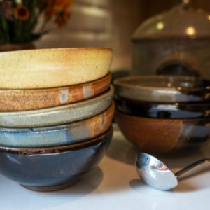 a horizontal framed photograph. a close up of two stacks of clay coyote chili bowls resting on a white kitchen countertop. behind them out of focus on the kitchens oven range, is a clay coyote flameware dutch oven with lid on. The stack of bowls closest to the left is 5 bowls high all nested. the top bowl is glazed in yellow salt, followed by joes blue, mint chip in the middle, another joes blue and a dark glaze on the bottom bowl. directly next to this stack of bowls is a silver colored soup ladle. directly behind the soup ladle is the second stack of chili bowls, three high. the top of this stack of chili bowls is glazed in mint chip, followed by a dark glaze, possibly zappa, then a joes blue on the bottom.