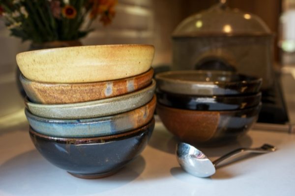 a horizontal framed photograph. a close up of two stacks of clay coyote chili bowls resting on a white kitchen countertop. behind them out of focus on the kitchens oven range, is a clay coyote flameware dutch oven with lid on. The stack of bowls closest to the left is 5 bowls high all nested. the top bowl is glazed in yellow salt, followed by joes blue, mint chip in the middle, another joes blue and a dark glaze on the bottom bowl. directly next to this stack of bowls is a silver colored soup ladle. directly behind the soup ladle is the second stack of chili bowls, three high. the top of this stack of chili bowls is glazed in mint chip, followed by a dark glaze, possibly zappa, then a joes blue on the bottom.
