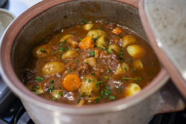 a horizontal framed picture. a clay coyote flameware dutch oven is filled with brown stew, the lid of the dutch oven is partially visible on the upper right. the lid is resting partially on the right "side" of the dutch oven. the dutch oven itself is slightly out of focus as the close up is centering on the stew contained within. the stew contains yellow potatoes, sliced carrot coins, meat and fresh green herbs sprinkled on top.