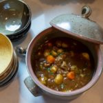 a horizontally framed photograph with a top down perspective shows a clay coyote flameware dutch oven filled with a brown hearty stew. to the left of the dutch oven is two stacks of clay coyote bowls. the top bowl on the lower stack is in yellow salt, the bowl on top of the stack above it is glazed in mint chip. the lid to the dutch oven is off, and resting on the upper left portion of the dutch ovens bases lip. between the dutch oven and the two stack of bowls, a small silver serving ladles head is visible, the handle is hidden. the dutch oven, bowls and ladle are all resting on a white kitchen countertop. there is a mix of outside white light and the kitchens warm yellow light.