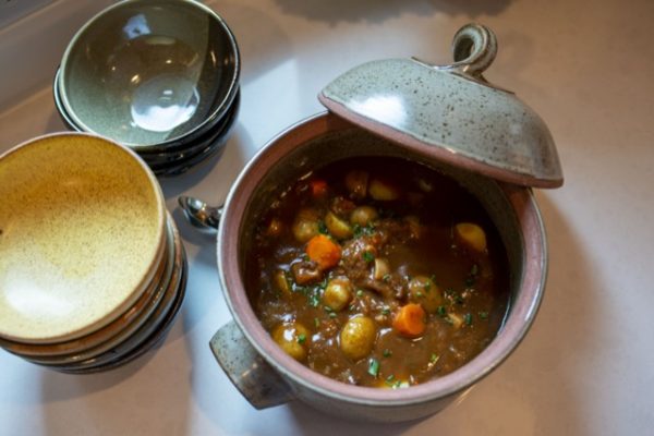 a horizontally framed photograph with a top down perspective shows a clay coyote flameware dutch oven filled with a brown hearty stew. to the left of the dutch oven is two stacks of clay coyote bowls. the top bowl on the lower stack is in yellow salt, the bowl on top of the stack above it is glazed in mint chip. the lid to the dutch oven is off, and resting on the upper left portion of the dutch ovens bases lip. between the dutch oven and the two stack of bowls, a small silver serving ladles head is visible, the handle is hidden. the dutch oven, bowls and ladle are all resting on a white kitchen countertop. there is a mix of outside white light and the kitchens warm yellow light.