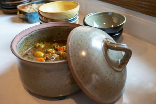 a horizontal orientated photograph of a clay coyote flameware dutch oven and two stacks of clay coyote chili bowls behind it. the dutch oven is filled with a hearty brown stew. the stew has meat, yellow potatoes, carrot slices and freshly cut herbs on top, all swimming in a thick brown stew broth. the lid of the dutch oven is resting on the "right" side of the dutch oven, with the lid standing diagonally leaning against the dutch oven base. behind the dutch oven is two stacks of clay coyote chili bowls. the top bowl of the left stack is in yellow salt glaze, and the bowl below that one is in joes blue. the top bowl of the right stack of bowls is glazed in mint chip, the two bowls below it are hard to ID the glaze. behind and to the left of the left stack of bowls, a partial view of a clay coyote baking dish in joes blue glaze is seen, it is filled with something but is out of focus. all of this is on a white kitchen countertop. there is natural white light coming from a nearby but unseen window, this is mixing with the warm yellow light of the kitchen.