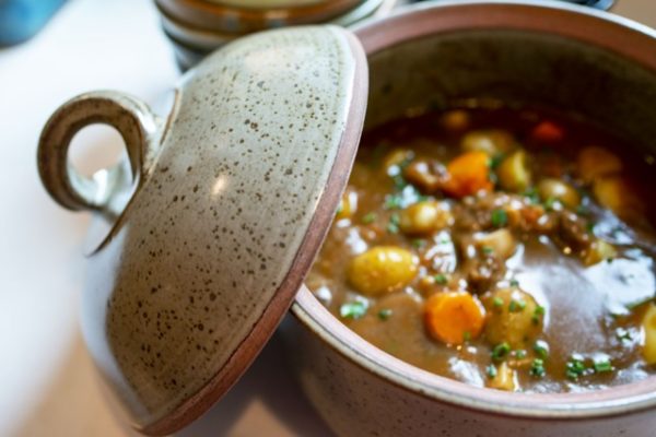 a horizontal orientated photograph. a clay coyote dutch oven is pictured from a lower then top down angle. The dutch oven is filled with a hearty meat and vegetable stew. the stew is rich brown with carrots, potatoes and beef all visible. the chef has sprinkled on fresh herbs over the top. the lid to the dutch oven is resting on the side of the dutch oven base, towards the left of the photo. the dutch oven is in coyote grey with dark speckling through out the glaze. behind the dutch ovens resting lid a stack of clay coyote bowls is just visible in frame, out of focus. there is a mix of white natural light and warm yellow light from the kitchen.