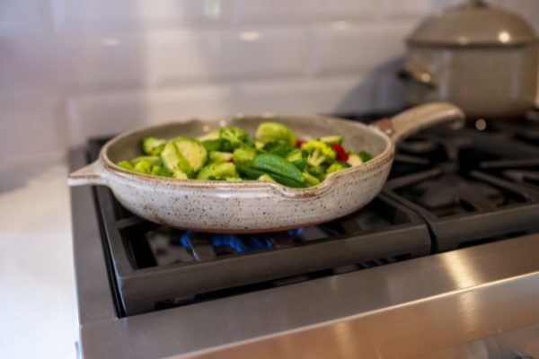 a horizontal orientated photograph of a clay coyote flameware large skillet. The skillet is in coyote grey with dark speckles through out the glaze. The large skillet is sitting on the iron grate of a lit gas kitchen range burner. there is a low small blue flame visible underneath the large skillet. The photograph was taken at a low angle, allowing for the side of the skillet to be visible, while still be able to see the entire rim of the skillet. The main handle is pointed towards the right side of the photograph, while on the exact opposite side the secondary smaller triangular handle is pointing to the left of the photograph. there are cut up vegetables cooking in the skillet, they include halved brussel sprouts, whole green pea pods, broccoli florets and sliced red bell pepper. The