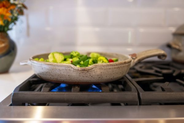 a horizontal orientated photograph shows a clay coyote flameware large skillet sitting on the iron grate of a lit gas burner in a home kitchen. There is blue flame visible underneath the large skillet. The photograph is taken from a low angle, slightly higher then the large skillet itself. Inside the skillet is various vegetables including halved brussel sprouts, whole pea pods, broccoli florets and sliced red bell peppers. The handle of the pan is pointing towards the right side of the frame. directly opposite the large handle is a smaller triangular handle facing to the left. partially cut out of picture on the left side is a clay coyote vase, and partially cut out on the right side is a clay coyote dutch oven, the same color (coyote grey) as the large skillet. The ovens backsplash is white tile.