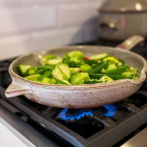 a vertical orientated photo shows a close up of a clay coyote large flameware skillet filled with green vegetables on a lit gas burner. Behind the skillet towards the upper right of the photograph is a clay coyote flameware dutch oven with lid on. It is also on the stop top, and is slightly out of focus. The vegetables in the skillet include: halved brussel sprouts, whole pea pods, broccoli florets and slices of red bell pepper. They all have a light sheen to them.