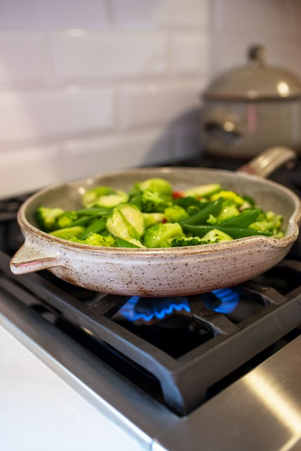 a vertical orientated photo shows a close up of a clay coyote large flameware skillet filled with green vegetables on a lit gas burner. Behind the skillet towards the upper right of the photograph is a clay coyote flameware dutch oven with lid on. It is also on the stop top, and is slightly out of focus. The vegetables in the skillet include: halved brussel sprouts, whole pea pods, broccoli florets and slices of red bell pepper. They all have a light sheen to them.