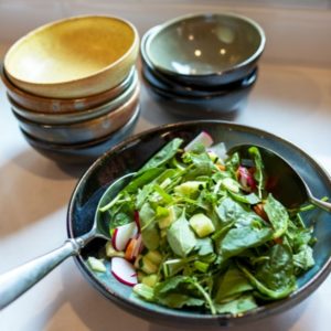 a vertical orientated shot of a clay coyote shallow serving bowl filled with a undressed spinach salad. in the salad are cut up radishes cucumbers and carrots. There are salad serving utensils on either side of the salad. Behind the salad is two stacks of clay coyote chili bowls. The left stack has 5 bowls and vary in glaze. The glazes descending from the top are, yellow salt, joes blue, mint chip, joes blue and zappa. The second stack of bowls consists of 3 bowls stacked the same way. The top of the 3 stack is in mint chip glaze. All the bowls are sitting on a white kitchen countertop and are seen at a 3/4th view. at the top of the shot a wooden window frame is seen out of focus. There is white natural light coming through the window, which is mostly overpower the soft warm yellow light of the kitchen.