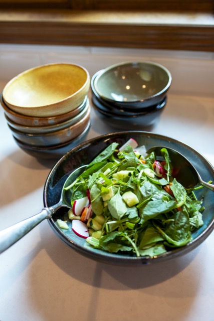 a vertical orientated shot of a clay coyote shallow serving bowl filled with a undressed spinach salad. in the salad are cut up radishes cucumbers and carrots. There are salad serving utensils on either side of the salad. Behind the salad is two stacks of clay coyote chili bowls. The left stack has 5 bowls and vary in glaze. The glazes descending from the top are, yellow salt, joes blue, mint chip, joes blue and zappa. The second stack of bowls consists of 3 bowls stacked the same way. The top of the 3 stack is in mint chip glaze. All the bowls are sitting on a white kitchen countertop and are seen at a 3/4th view. at the top of the shot a wooden window frame is seen out of focus. There is white natural light coming through the window, which is mostly overpower the soft warm yellow light of the kitchen.