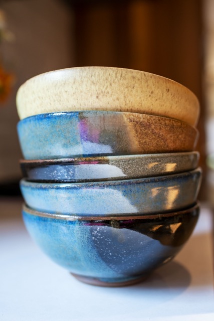 a stack of 5 clay coyote bowls are seen nested into one another from table level. The bowls vary in glaze color and have a yellow salt bowl on the top followed by a joes blue, mint chip, and zappa. The bowl stack is sitting on a white countertop with white light reflecting off of the shiny parts of the glazes. The yellow salt bowl on the top is matte, and the brown part of the joes blue bowl directly under it is also matte.