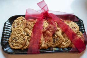 a clay coyote large tray in midnight garden glaze is filled with baked cookies. The shot is horizontal and at a 3/4 view. The cookies are stacked in an alternating manner like fish scales. there are around 13 light brown cookies, with large chunks of dark add ins visable. a red mesh ribbon bow is wrapped around the tray , with the lower right part of the tray and middle left of the tray being wrapped in the ribbon.