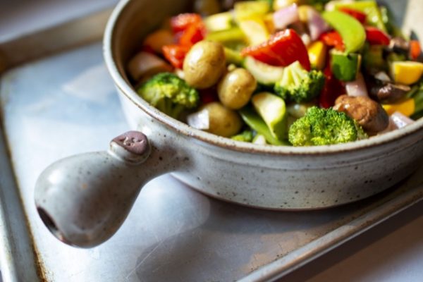 a horizontal orientated photo. a clay coyote flameware grill basket in coyote grey is pictured from high side view. The handle of the grill basket is pointed to the lower left side of the picture. the grill basket is sitting on a silver yellow backing sheet, which itself is sitting on a white kitchen counter top. The grill basket is filled with a very colorful assortment of vegetables. They include: whole yellow baby potatoes, broccoli florets, zucchini, purple onion slices, red and green pepper slices and yellow summer squash. It is lit with white natural light, which catches on the handle and baking sheet