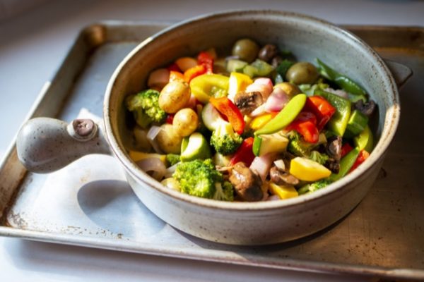 a horizontal orientated photo. A clay coyote flameware grill basket is filled with colorful cut up vegetables. The entire grill basket is sitting on a silver baking sheet, which is in turn sitting on a white kitchen countertop. The grill basket's bulbous handle is pointed to the left side of the picture. The cut up vegetables include: red and green bell peppers, broccoli florets, whole pea pods, whole yellow baby potatoes, purple onion, yellow summer squash and mushroom slices. The colorful mix is partially lit by a direct sun beam, allowing the fresh vegetables colors to really pop.