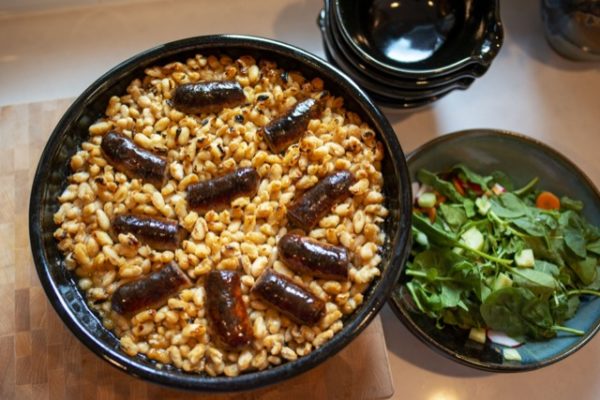 on the center to center left of the picture is a clay coyote special edition "cassoulet confessions" cassole filled with white/yellow beans and sausage. The rim of the cassole is midnight black. The beans and sausage pieces show evidence of oven roasting. The sausage looks plump and glistening. To the right of the picture is a spinach salad with cut up radish, carrots and cucumber. The salad is in a clay coyote zappa glaze bowl. On the upper right of the shot, 1/4 cut off out of frame, is a stack of 4 empty clay coyote cassole bowls in midnight black glaze also. the special edition cassole is sitting on a butchers block, while the salad and stack of bowls is on the kitchen countertop. There is warm kitchen light mixing with white light outside.