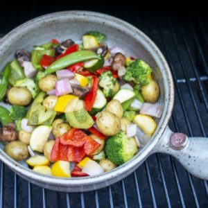 a clay coyote flameware grill basket is viewed from the top down. The grill basket is grey with darker speckles through out the glaze. The top down view shows that there are cut up vegetables and baby potatoes in the pan. The vegetables are a mix of bright greens, yellows, reds and some light purple mixed in with cut up onion. The grill basket is sitting on top of a grill grate, which has black bars running vertical in the frame.