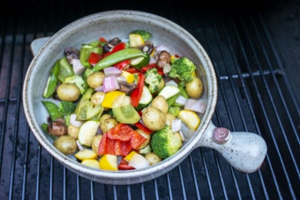 a clay coyote flameware grill basket is viewed from the top down. The grill basket is grey with darker speckles through out the glaze. The top down view shows that there are cut up vegetables and baby potatoes in the pan. The vegetables are a mix of bright greens, yellows, reds and some light purple mixed in with cut up onion. The grill basket is sitting on top of a grill grate, which has black bars running vertical in the frame.