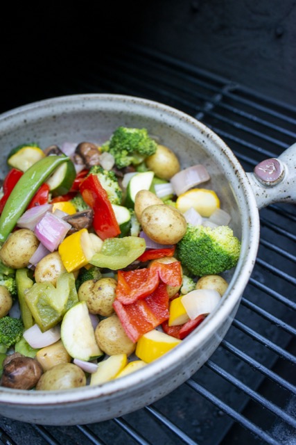 a clay coyote flameware grill basket is seen from 3/4 view. The grill basket is sitting on a grill with the black grill bars visible under the basket. The grill basket is filled with brightly colored cut up vegetables and baby yellow potatoes. The grill basket is partially out of frame on the left side of the shot, and the handle on the right side is half visible. The picture is in soft white daylight.