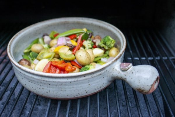 a clay coyote flameware grill basket is seen from 3/4 view. The grill basket is sitting on a grill with the black grill bars running vertical, visible under the basket. The grill basket is filled with brightly colored cut up vegetables and baby yellow potatoes. the picture is in soft white daylight.