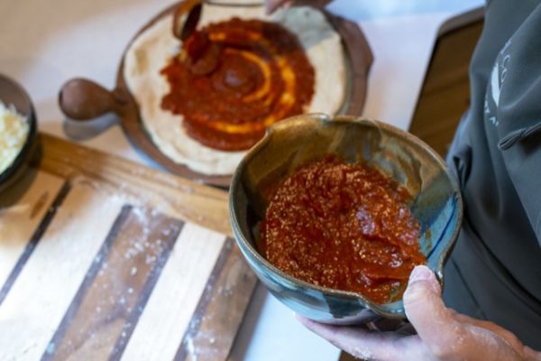 a horizontally framed photograph shows a near overhead view of a clay coyote whiskey bowl that is halfway filled with pizza sauce. the pizza chef is holding the whiskey bowl in their left hand, nearer to the camera. with their right hand they are using a small ladle to spread pizza sauce over the raw pizza dough that is resting on a clay coyote pizza stone. the whiskey bowl is glazed in joes blue. the pizza stone is resting on a white kitchen counter top and is slightly out of focus. also on the kitchen countertop is a flour dusted wooden cutting board. the wooden cutting board has alternating wide stripes of light and darker wood. between each stripe there is a thin stripe of very dark wood.