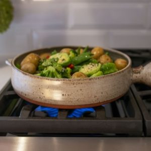 a horizontal framed picture of a clay coyote flameware cazuela cooking vegetables on a kitchen stove top range. the picture is taken at a slightly higher then side angle viewpoint allowing the viewer to see the vegetables cooking within. The vegetables include: yellow whole baby potatoes, brussel sprouts halved, broccoli florets, and whole pea pods. There are a couple strips of red bell pepper mixed in. All the vegetables in the cazuela have a light oil sheen to them and cracked pepper is visible. There is blue flame visible under the cazuela. Behind and to the upper left of the photo is a clay coyote small vase in feather glaze. The cazuela is in coyote grey, with small darker speckles through out. on the left of the picture a small strip of white countertop is visible and is what the vase is resting on.