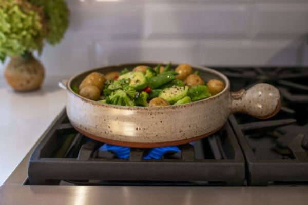 a horizontal framed picture of a clay coyote flameware cazuela cooking vegetables on a kitchen stove top range. the picture is taken at a slightly higher then side angle viewpoint allowing the viewer to see the vegetables cooking within. The vegetables include: yellow whole baby potatoes, brussel sprouts halved, broccoli florets, and whole pea pods. There are a couple strips of red bell pepper mixed in. All the vegetables in the cazuela have a light oil sheen to them and cracked pepper is visible. There is blue flame visible under the cazuela. Behind and to the upper left of the photo is a clay coyote small vase in feather glaze. The cazuela is in coyote grey, with small darker speckles through out. on the left of the picture a small strip of white countertop is visible and is what the vase is resting on.