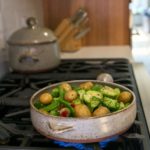 a vertical orientated shot. a clay coyote flameware cazuela sitting on a lit gas burner, filled with cooking vegetables. The vegetables include yellow baby potatoes, broccoli florets, whole pea pods, red bell pepper slices and halved brussel sprouts. The veggies have a light sheen of oil on them and cracked pepper visible through out. The cazuela handle is pointed away from the viewer, and the small secondary triangle handle is visible. The cazuela is grey with darker speckles through out the glaze. The very bottom of the cazuela is unglazed bare pottery. Behind and to the left of the frame is a slightly out of focus clay coyote dutch oven. The lid is on. further behind the dutch oven and out of focus is a knife block sitting on white kitchen counter top.