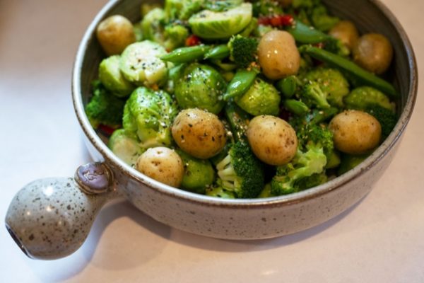 a horizontal show picture at 3/4 view. a clay coyote flameware cazuela is seen filled with cooked broccoli florets, yellow baby potatoes, halved brussel sprouts and whole pea pods. The greens are vibrant, and there is a light oil visible. The veggies have been seasoned with cracked pepper. The handle of the cazuela is pointed towards the lower left side corner. The cazuela is in coyote grey with darker spots visible through out the glaze. There is white daylight viable on the lower right. The cazuela is sitting on a white kitchen countertop.