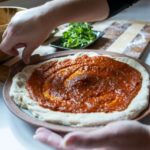 a horizontally framed photograph shows a clay coyote pizza stone that is being used to make a home made pizza. the pizza dough has been placed on the stone and sauce has been spread over it. the chef making it is currently in process of pressing their right thumb into the crust section of the dough to give the crust more texture/shape. their left hand is palm up, their right hand has their thumb down and it is very close to the dough, and it is visible where they had pressed their thumb into the crust. behind the pizza stone is a flour dusted cutting board. the cutting board has alternating wide stripes of light and medium brown colored wood. each wide stripe is divided by a thin stripe of dark brown colored wood. there is a clay coyote small tray with a bunch of fresh herbs in it. near the chefs right hand and in the upper left corner of the photograph is a clay coyote deep salad bowl glazed in feather. due to the camera angle it isn't possible to see what is inside. the lighting is natural white light from a nearby unphotographed window.