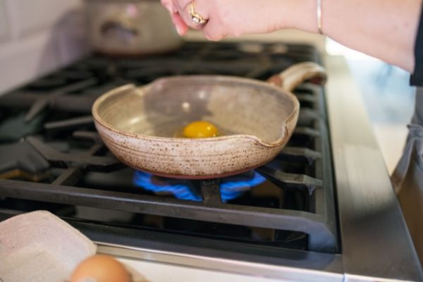 a clay coyote flameware small skillet is center frame. It is grey with speckles through out the glaze. A woman's hands are visible coming from the right of the shot, she is just about to crack a 2nd egg into the small skillet. The skillet sits on top of a lit gas burner on a kitchen range. In the foreground just barely in the frame is the edge of a grey egg carton with the top of one brown egg barely peaking into frame. Behind and to the left of the small skillet is a clay coyote dutch oven, the top of the dutch oven is not in frame, and it is only visible out of focus and from the handle and below.