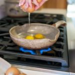 a clay coyote flameware small skillet sits in the middle of the frame. The skillet is grey with speckled through out the glaze. A woman's hands are seen cracking the second egg of two into the pan, the brown egg shell is cracking open and egg is still falling from the egg shell into the pan. In the pan two yokes are visible. The pan is on a lit gas burner with blue flame visible beneath it. In the foreground to the lower left of the shot, two brown eggs are sitting in a grey egg carton. Only the last 4 egg compartments are seen, the rest is out of shot. Behind the clay coyote flameware small skillet towards the upper left of the shot is a slightly out of focus clay coyote dutch oven with lid on. it is the same color as the skillet.