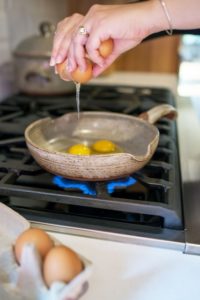 a clay coyote flameware small skillet sits in the middle of the frame. The skillet is grey with speckled through out the glaze. A woman's hands are seen cracking the second egg of two into the pan, the brown egg shell is cracking open and egg is still falling from the egg shell into the pan. In the pan two yokes are visible. The pan is on a lit gas burner with blue flame visible beneath it. In the foreground to the lower left of the shot, two brown eggs are sitting in a grey egg carton. Only the last 4 egg compartments are seen, the rest is out of shot. Behind the clay coyote flameware small skillet towards the upper left of the shot is a slightly out of focus clay coyote dutch oven with lid on. it is the same color as the skillet.