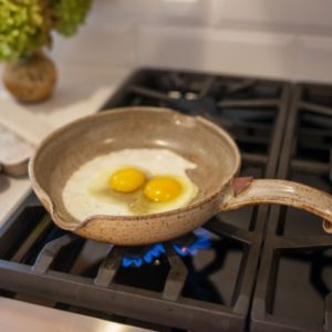center frame is a clay coyote flameware small skillet with two freshly cracked eggs in the middle of the pan. The skillet has the handle facing to the right of the frame. The skillet is grey with dark speckles through out the glaze.They yokes look raw and the whites of the eggs are just starting to turn from clear to white, with a small bit of clear eggs still around the two yokes in the center of the small skillet. The gas burner is lit, with small blue flames visible