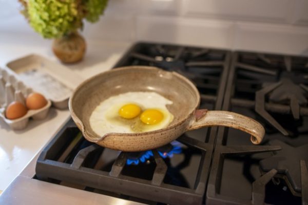 center frame is a clay coyote flameware small skillet with two freshly cracked eggs in the middle of the pan. The skillet has the handle facing to the right of the frame. The skillet is grey with dark speckles through out the glaze.They yokes look raw and the whites of the eggs are just starting to turn from clear to white, with a small bit of clear eggs still around the two yokes in the center of the small skillet. The gas burner is lit, with small blue flames visible