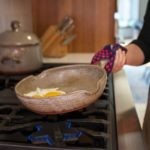 a woman in a dark shirt and grey apron is holding a red and black checkered kitchen hot pad around the handle of a clay coyote flameware small skillet. There are two eggs inside the small pan which is held at a slight angle over a lit gas burner. In the background, also on the stove top is a clay coyote flameware dutch oven with lid on, behind that is a knife block slightly out of focus.