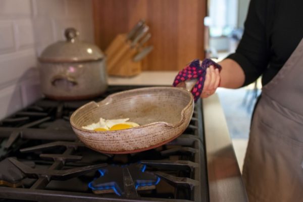 a woman in a dark shirt and grey apron is holding a red and black checkered kitchen hot pad around the handle of a clay coyote flameware small skillet. There are two eggs inside the small pan which is held at a slight angle over a lit gas burner. In the background, also on the stove top is a clay coyote flameware dutch oven with lid on, behind that is a knife block slightly out of focus.