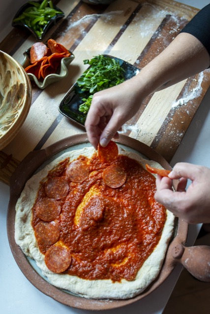 a vertically framed photograph shows an overhead view of a clay coyote pizza stone that is being used currently to make a home made fresh pizza. the pizza dough is raw, with red pizza sauce covering most of it. the chef is currently placing down a layer of pepperonis. their right hand is coming in from the top right corner and their left hand is just coming into frame about 2/3rds of the way down the right side of the photograph. both of the chef's hands are holding onto a pepperoni slice. they are in process of laying down a layer of pepperonis on top of the pizza sauce. 6 pepperonis in a half circle on the outer edge of the pizza sauce zone have already been laid down. on the upper left edge of the photograph a clay coyote deep salad bowl can be partially seen. it is glazed in feather. slightly to the upper right of the bowl are two clay coyote little dippers, one is filled with pepperonis and the other is filled with sliced green peppers. at the "12 o'clock" position from the pizza stone is a clay coyote small tray glazed in midnight garden. it is holding a bunch of fresh green herbs.