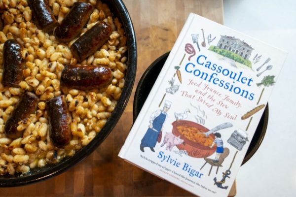 Special edition Clay Coyote Cassole, with a black rim filled with cooked white beans and sausage. The cassole is on the left of the picture, on the right is "Cassoulet Confessions" a white book. Both the book and the Cassole are resting on a wooden cutting board