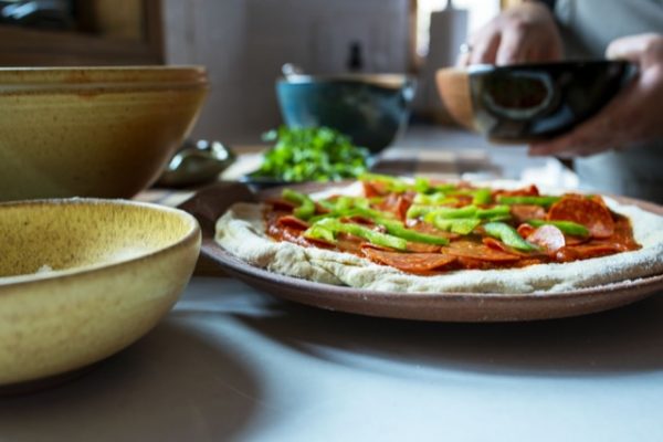 a horizontally framed photograph show a clay coyote pizza stone being used to make a fresh raw pizza. the pizza stones cooking surface is almost completely covered by the raw dough. only the edges of the pizza stone are visible due to the pizza resting on it. the outside edge and secondary handle (the primary bulbous handle is not visible, and is cut out of frame on the right side) are unglazed and show off the natural reddish brown of the clay. the raw pizza has sauce, a layer of pepperonis, a layer of green peppers and the chef is currently holding a clay coyote chili bowl glazed in mocha swirl, with one hand reaching into it. near the bottom left of the of the photograph is a clay coyote chili bowl, but the camera angle prevents from seeing inside, as it is just a bit above a side on view. it is glazed in yellow salt. directly behind the yellow salt soup/chili bowl is a deep salad bowl glazed in feather. it also is a side view due to the camera angle. much further down the kitchen counter top everything is sitting on, and out of focus is a clay coyote whiskey bowl, it is glazed in joes blue. it is resting on a wooden cutting board that has wide light and medium colored wood stripes. dividing each of these wide stripes is a thin dark wood stripe. there is an out of focus bunch of fresh herbs sitting on the cutting board.