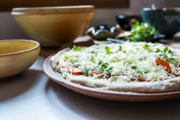 a horizontally framed photograph shows a clay coyote pizza stone that has a fresh raw home made pizza resting on it, a clay coyote deep salad bowl glazed in feather and a clay coyote chili bowl glazed in yellow salt. the photograph is taken just a couple inches above the counter top height on which they are all resting. due to this angle it is not possible to see what is in the chili bowl or the salad bowl, as they are seen from just about a side on angle. the pizza stones cooking surface is almost completely covered by the raw pizza. the pizza crust has been floured or dusted with fine corn meal, there are pepperoni slices peaking out of the shredded cheese from a few spots. the pizza is covered with white shredded cheese. there are fresh herbs sprinkled on top of the pizza also. in the background out of focus there is a clay coyote whiskey bowl, possibly another chili bowl and a little dipper.