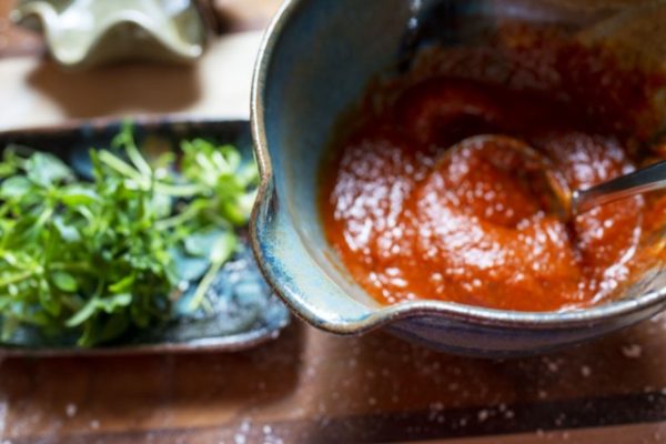 a horizontally framed photograph shows a close up almost overhead view of a clay coyote whiskey bowl and a clay coyote small tray without handles. the small tray is filled with fresh herbs. the whiskey bowl is filled with pizza sauce. the whiskey bowl is glazed in joes blue, and is photographed from so close that not all of the bowl can be seen in the photograph. the whiskey bowl with pizza sauce has a small ladle with a circular spoon head resting inside the bowl. in the very upper left corner of the photograph is an out of focus and half out of frame clay coyote little dipper in mint chip. all of these pieces are resting on a wooden cutting board. the wooden cutting board has wide stripes of light and medium colored wood, which is divided by thin stripes of dark wood. there are flour particles visible on the wooden cutting board. the focus of the photograph is on the lip edge of the whiskey bowl, with everything else in the photograph being slightly out of focus. the photograph is lit by natural white light.