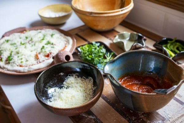 a horizontally framed photograph shows various clay coyote serving and cooking pieces that have been used to make a fresh home made pizza. starting from the left of the photograph moving right there are: a clay coyote pizza stone, two clay coyote chili bowls, a deep salad bowl, a small tray (without handles), a clay coyote whiskey bowl, a little dipper and finally another little dipper. the pizza stone has a whole fresh made raw pizza resting on its cooking surface. it has sauce, pepperonis, green peppers, shredded white cheese, and fresh herbs. the first chili bowl is glazed in yellow salt, and has corn meal in it. the next chili bowl has shredded white cheese. the next serving piece from left to right is a clay coyote small tray without handles, it is filled with a bunch of fresh green herbs. the small tray has dark glaze, and is possibly glazed in zappa. the deep salad bowl is angled at a way it isn't possible to see what is inside of it, but it is glazed in feather. the next piece while moving left to right would be a clay coyote whiskey bowl, glazed in joes blue. it has red pizza sauce, and a small silver ladle resting inside. finally comes the two little dippers, the first is in the start pattern, and is glazed in mint chip, there doesn't appear to be anything inside of it. finally there is a square style little dipper, it has sliced green peppers inside of it. all of this but the chili bowl in yellow salt are resting on a wooden cutting board. the cutting board has wide stripes of light and medium colored wood, with small thin stripes of dark wood dividing them. the chili bowl in yellow salt is not resting on the cutting board, but rather next to it on a white kitchen counter top. the lighting in the picture is white natural light, coming from a nearby but unseen window.