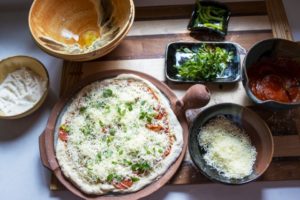 a horizontally framed photograph shows multiple clay coyote pottery pieces being used to make a fresh pizza. working left to right there is a clay coyote chili bowl glazed in yellow salt, that contains finely ground corn meal. to the upper right of that bowl is a clay coyote deep salad bowl glazed in feather. it has had some liquid recently used in it, as there is nothing in the bowl currently but there is a liquid shine inside the bowl. below that is a clay coyote pizza stone, which has the fully assembled but still raw pizza on its cooking surface. to the right of the pizza stone/pizza is another clay coyote chili bowl, this one is filled with shredded white cheese. above that or at the "2 o'clock" position relative to the pizza stone, is a clay coyote small tray with out handles. it has fresh herbs inside it. directly above that piece is a clay coyote little dipper (the square style) with sliced green pepper pieces inside of it. finally to the right of the small tray with the herbs is a clay coyote whiskey bowl filled with the pizza sauce. all the pieces listed except for the first (the chili bowl with cornmeal) are resting on a wooden cutting board. the wooden cutting board has wide stripes of light and medium colored wood that is divided by thin stripes of dark wood. the cutting board and the chili bowl with the corn meal are resting on a white kitchen counter top.