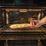 a horizontally framed photograph shows an arm placing a clay coyote pizza stone with a fresh raw pizza into an oven. the arm is coming from the right side of the photograph, with only the fore arm and hand visible. the hand is palm up using the primary bulbous handle of the pizza stone to place it onto the top rack (of two). the pizza is clearly raw, as the dough is still lightly colored and the shredding cheese is still not melted. the chef is also using their bare hands to place it in, indicating that it is not hot yet. the oven is lit by two long lights on either vertical wall.