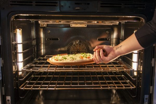a horizontally framed photograph shows an arm placing a clay coyote pizza stone with a fresh raw pizza into an oven. the arm is coming from the right side of the photograph, with only the fore arm and hand visible. the hand is palm up using the primary bulbous handle of the pizza stone to place it onto the top rack (of two). the pizza is clearly raw, as the dough is still lightly colored and the shredding cheese is still not melted. the chef is also using their bare hands to place it in, indicating that it is not hot yet. the oven is lit by two long lights on either vertical wall.