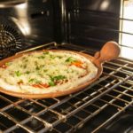 a horizontally framed photograph shows a clay coyote pizza stone in use. it is on the top rack of an oven, cooking a fresh pizza. the pizza is freshly made and all the ingredients are still raw. the pizza stones cooking surface is completely covered by the baking pizza, only its non glazed edge and primary bulbous and secondary handles are visible, showing off the natural color of the clay. the pizza cooking on the stone looks to have corn meal on the crust, with plenty of white cheese layered on top of pepperonis. on top of the cheese layer there is small green herbs cut up and sprinkled over the pizza. the light in the oven in white, and there is warm yellow light coming from somewhere unseen. there are two racks in the oven. it is a convection oven, as there is a visible fan/blower in the back.