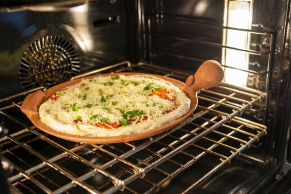 a horizontally framed photograph shows a clay coyote pizza stone in use. it is on the top rack of an oven, cooking a fresh pizza. the pizza is freshly made and all the ingredients are still raw. the pizza stones cooking surface is completely covered by the baking pizza, only its non glazed edge and primary bulbous and secondary handles are visible, showing off the natural color of the clay. the pizza cooking on the stone looks to have corn meal on the crust, with plenty of white cheese layered on top of pepperonis. on top of the cheese layer there is small green herbs cut up and sprinkled over the pizza. the light in the oven in white, and there is warm yellow light coming from somewhere unseen. there are two racks in the oven. it is a convection oven, as there is a visible fan/blower in the back.