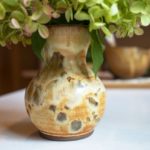 a horizontally framed photograph shows a close up of a clay coyote small vase glazed in feather. the vase has a bouquet of flowers in it, the flowers are mostly cut off out of frame, but the bit visible is bright green with hints of pink. the bouquet of flowers is larger then the vase. the vase is sitting on a white kitchen counter top. the photograph was taken at counter top height, giving the vase a "head on" view. in the background, to the right and further down the kitchen counter top is a clay coyote whiskey bowl glazed in joes blue. it is out of focus.