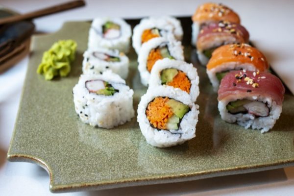 a horizontally framed photograph shows a clay coyote sushi plate in mint chip glaze being used to serve sushi. there are twelve pieces of sushi on the plate, and a dab of wasabi. starting from left to right on the plate: a dab of wasabi, 3 pieces of sushi, a row of five pieces then a row of 4 pieces of nigiri style, with alternating tuna and salmon pieces. the sushi plate is resting on a white kitchen countertop. the room is lit by clear white light. in the upper left corner of the photograph there is a small wooden spoon handle, but the rest is cut off and identifiable.