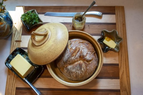 a horizontally framed photograph shows a overhead view of a clay coyote bread baker, 2 little dipper, small handleless tray and a glass jar of honey, all resting on a wooden cutting board. the bread baker is glazed in yellow salt, and has been used to bake a fresh loaf of bread. at the "12 oclock" position relative to the circular bread baker is a glass 6 sided jar of honey. the jar of honey has a silver metal spoon resting inside of it, with a large bread knife resting slightly behind it. at the "2 oclock" position relative to the bread baker is a clay coyote little dipper that has a knob of butter and some honey inside of it. the little dipper is glazed in mint chip. at the "8 to 9 oclock" position is a clay coyote small handleless tray with a block of butter on it, a butter knife is also on the tray. the tray is glazed in midnight garden. at the "10 oclock" position relative to the bread baker is another little dipper, this one is in the square style, it is filled with a fresh green herb. all of this is resting on a wooden cutting board. the wooden cutting board has alternating stripes of light and medium colored wood. these wide stripes are separated by thinner dark stripes of wood. in the very upper left corner there is a small sunbeam of light. directly next to the sunbeam is a clay coyote vase with a bouquet